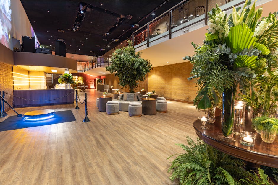 A spacious indoor venue with warm wooden flooring and a high ceiling, decorated with elegant floral arrangements. In the foreground, there is a tall, clear glass vase filled with white and green flowe