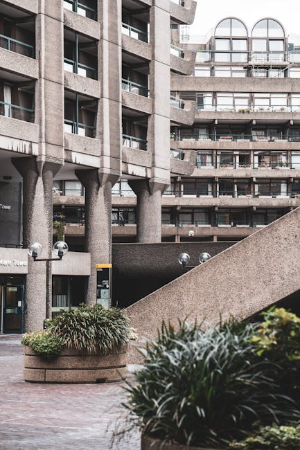 The image depicts the exterior of a large, modern concrete building with multiple floors and balconies, characteristic of a residential or commercial tower. In the foreground, there is a paved courtya