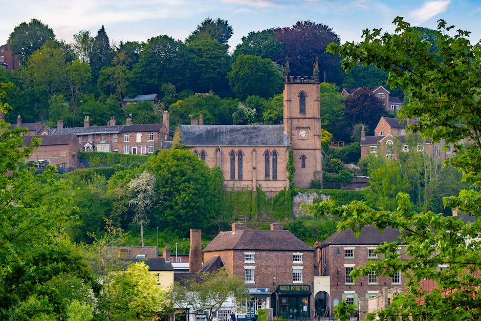 A scenic view of a small town with historic brick buildings and a prominent church with a tall, square tower, situated among lush green trees and foliage. The scene features residential houses on a hi