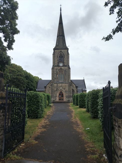The image depicts a historic stone church with a tall, pointed spire topped by a cross, situated at the end of a paved pathway leading through well-maintained green hedges. The church features a large