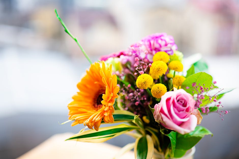 A vibrant floral arrangement featuring a bright orange gerbera daisy, a soft pink rose, and clusters of small yellow billy balls, complemented by pink and purple filler flowers and green foliage. The 