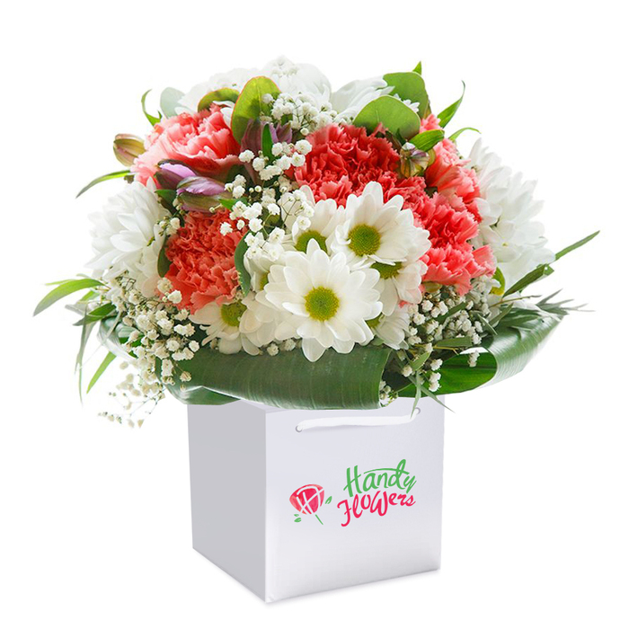 Poised upright within a pristine square white gift bag, this lush bouquet radiates with uplifting colour and artistry, as if gathered fresh from the vibrant flower stalls of St Luke's. A luxuriously full mix of soft coral and lively red carnations commands the centre, their ruffled petals bursting with depth and texture. Nestled between these statement blooms are clusters of crisp white daisies, their zesty lime-green centres illuminating the arrangement like sunlight trailing across the lawns of Finsbury Circus. Wisps of delicate baby's breath thread gracefully throughout, lending the piece a light, airy elegance that softens and connects each flower. Accents of deep green foliage-a mix of wide, glossy leaves and slender sprigs-form a verdant collar, grounding the bouquet in nature and hinting at the leafy avenues near City University. On the bouquet's left, hints of closed buds with subtle purple flushes add intrigue and progression, suggesting new beginnings. The white gift bag, with its strong white handles designed for convenience, is perfect for same-day local delivery right to a doorstep in St Luke's. Prominently displayed on the bag's face, the minimalist logo nods to the trusted local florist whose artistry is visible in every expertly placed stem. Imbued with a sense of occasion, this arrangement is the very embodiment of generosity and warmth-ideal for celebrations, anniversaries, or heartfelt gestures delivered fresh and beautiful across St Luke's vibrant community.