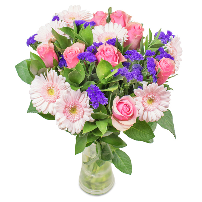 A vibrantly arranged bouquet captures the spirit of St Luke's, presented in a clear glass vase against a pristine white setting. The arrangement showcases about seven to nine pink roses-some with petals still curled tightly, others just beginning to reveal their classic spirals-reflecting the tender anticipation of new beginnings so often felt in the community along City Road. Intertwined among the roses, approximately five to seven pale pink gerbera daisies stand out, identifiable by their large, round blooms and slightly lighter, almost ivory, centers. Pops of vivid purple statice provide striking texture and depth, much like the colourful street art near Whitecross Street. Deep green foliage frames the bouquet, adding fullness and a lush, natural touch reminiscent of St Luke's pocket parks. The transparent vase, its water and stems clearly visible, suggests a freshly prepared gift ready for delivery, ideal for celebrating birthdays, anniversaries, or simply to bring warmth to an office or home nearby. This harmonious blend of roses, gerberas, and purple accents beautifully expresses joy, affection, and lively London charm.
